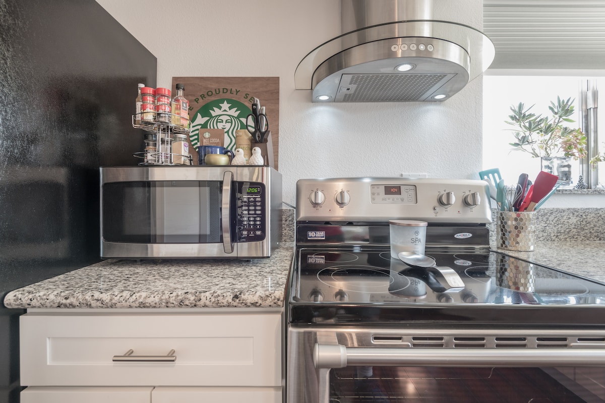 The kitchen area features modern appliances, including a stainless steel oven and microwave. A variety of utensils are displayed, alongside a selection of spices in clear containers on the counter. Natural light enters through a nearby window, illuminating the granite countertops.