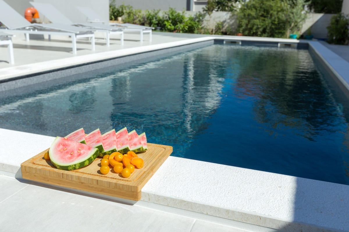 A wooden serving board holds slices of watermelon and yellow fruit, placed beside a swimming pool. The pool's clear water reflects sunlight, creating a bright atmosphere. Sun loungers are positioned nearby, surrounded by lush greenery.