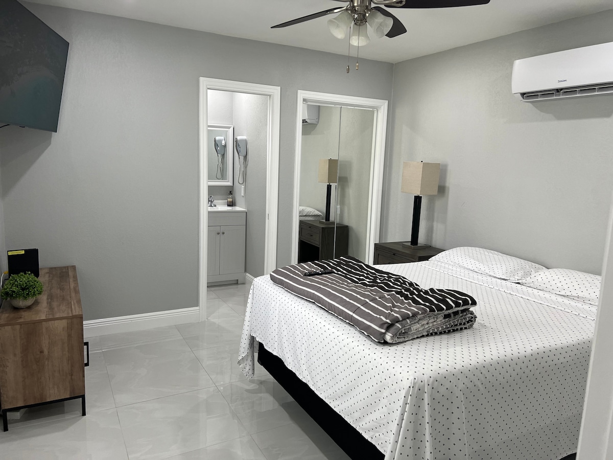 A spacious sleeping area is equipped with a large bed covered in a polka dot blanket. A ceiling fan is positioned above, and light streams in from a nearby window. Two bedside lamps flank the bed, and a television is mounted on the wall opposite the bed.