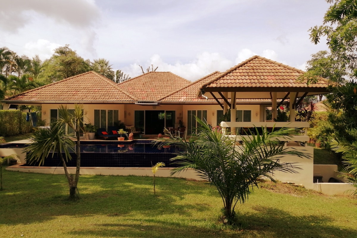 The spacious villa is framed by lush greenery, featuring a large infinity pool and an elevated gazebo. Sliding glass doors reveal views of the inviting patio area. The roof is tiled, and palm trees are visible in the foreground, enhancing the serene outdoor atmosphere.