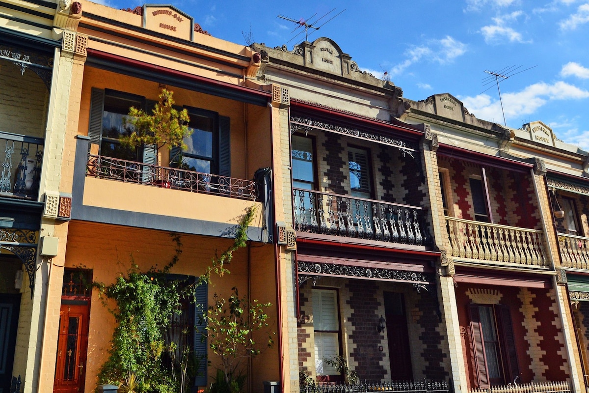 A row of Victorian terrace houses is displayed, featuring intricate architectural details. Each house showcases distinct facade colors and ornamental elements. Balconies with wrought iron railings are visible, along with decorative masonry work that highlights the elegant style of the early 1900s.