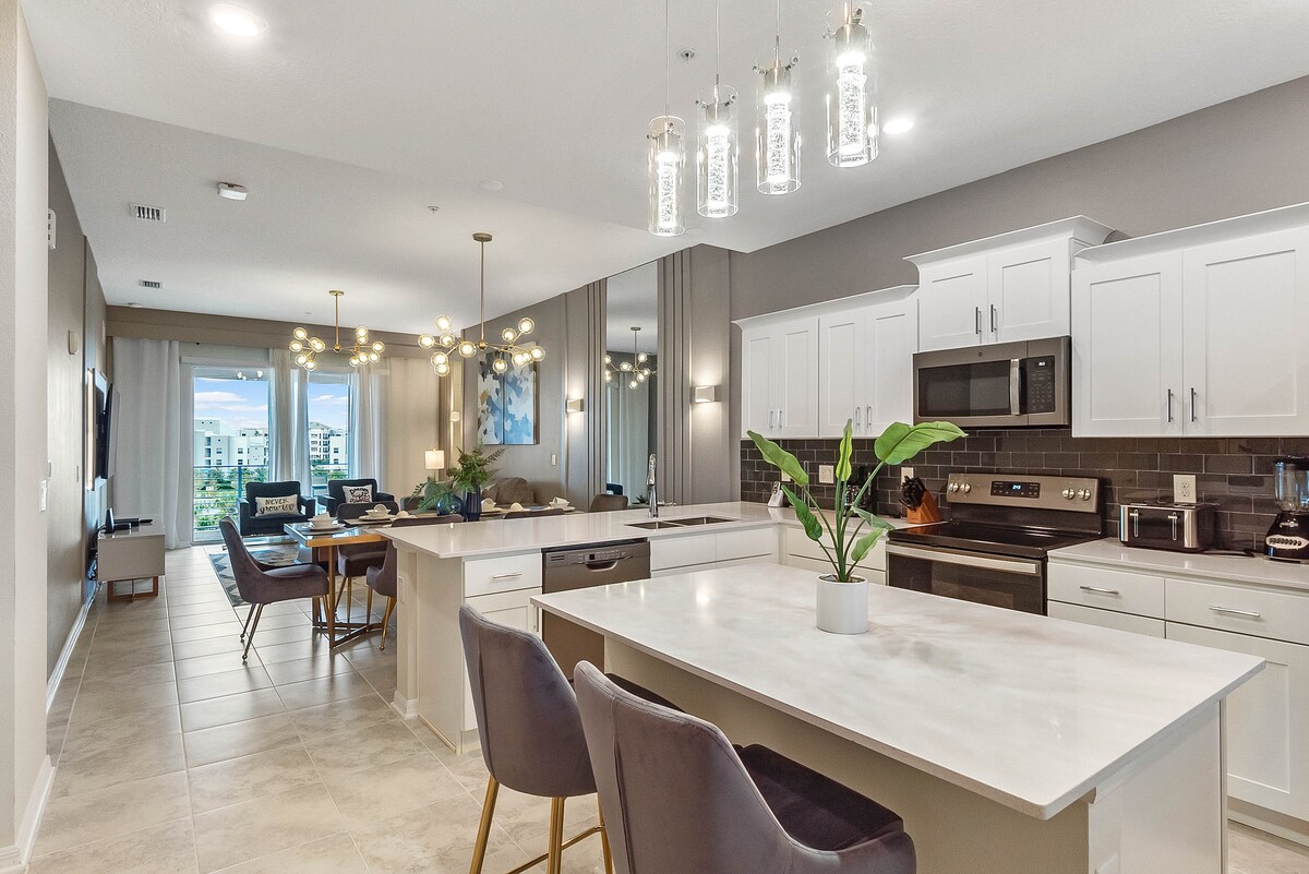 An open-concept kitchen and living area are featured, showcasing modern cabinetry and quartz countertops. The dining section includes a round table surrounded by four chairs. Statement lighting fixtures hang above the kitchen island, which is complete with seating. Large windows invite natural light and provide views of the outdoors.