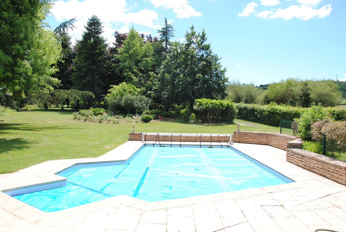An outdoor pool is showcased, surrounded by a spacious green lawn and various trees. The pool is covered with a light blue tarp, and a stone border is seen along the edge. The clear sky is visible, featuring a few scattered clouds.