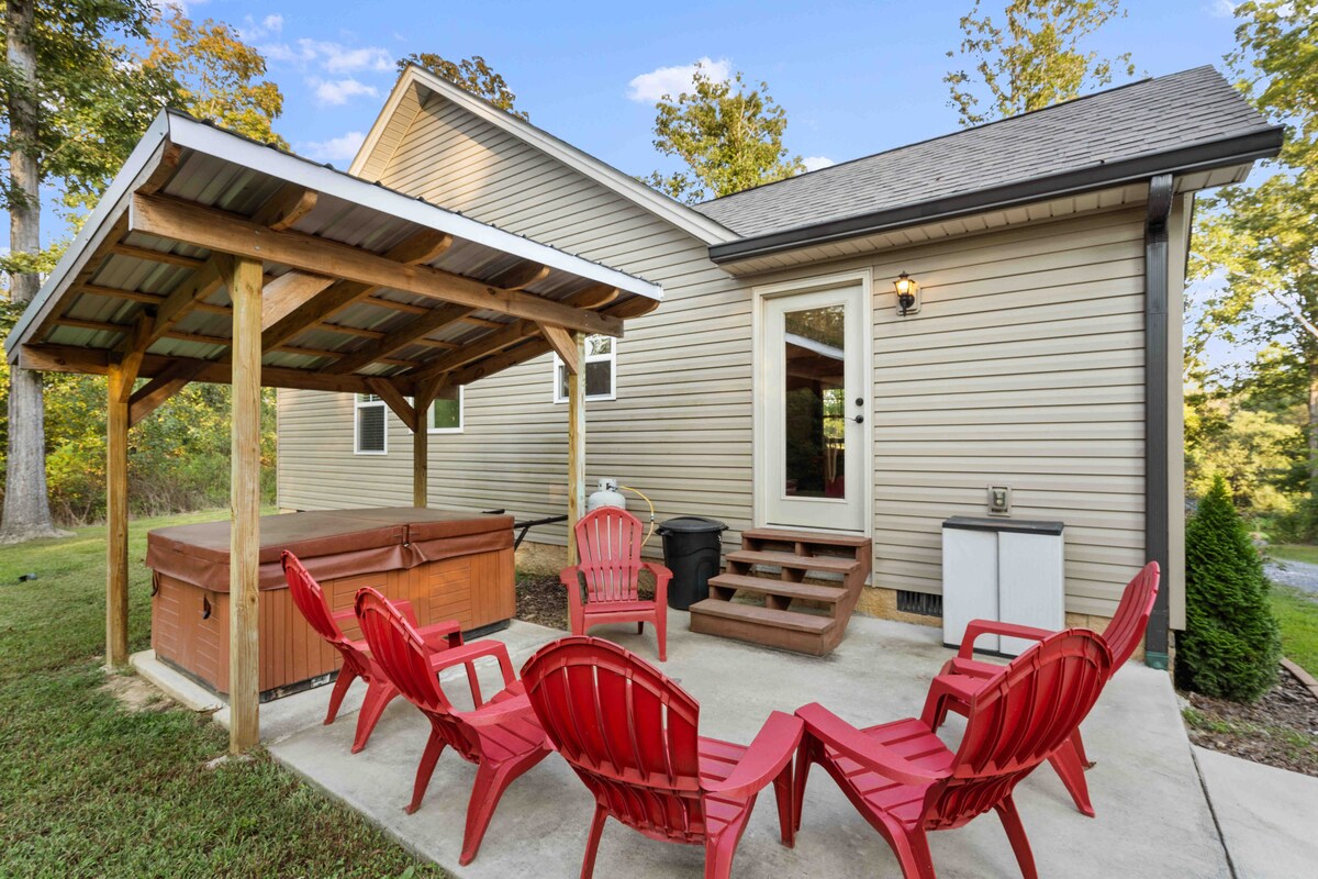 An outdoor patio area is presented, featuring a hot tub under a wooden shelter. Surrounding red Adirondack chairs offer a space for relaxation. Steps lead up to the entrance of the home, which is accentuated by a blend of natural surroundings and well-maintained grass.