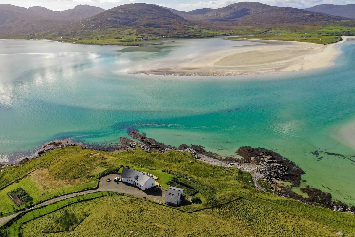 An aerial view captures the accommodation nestled on verdant land, surrounded by the turquoise waters of Luskentyre beach. The sandy shore and rolling hills create a serene landscape, highlighting the peaceful coastal environment of the Hebrides.