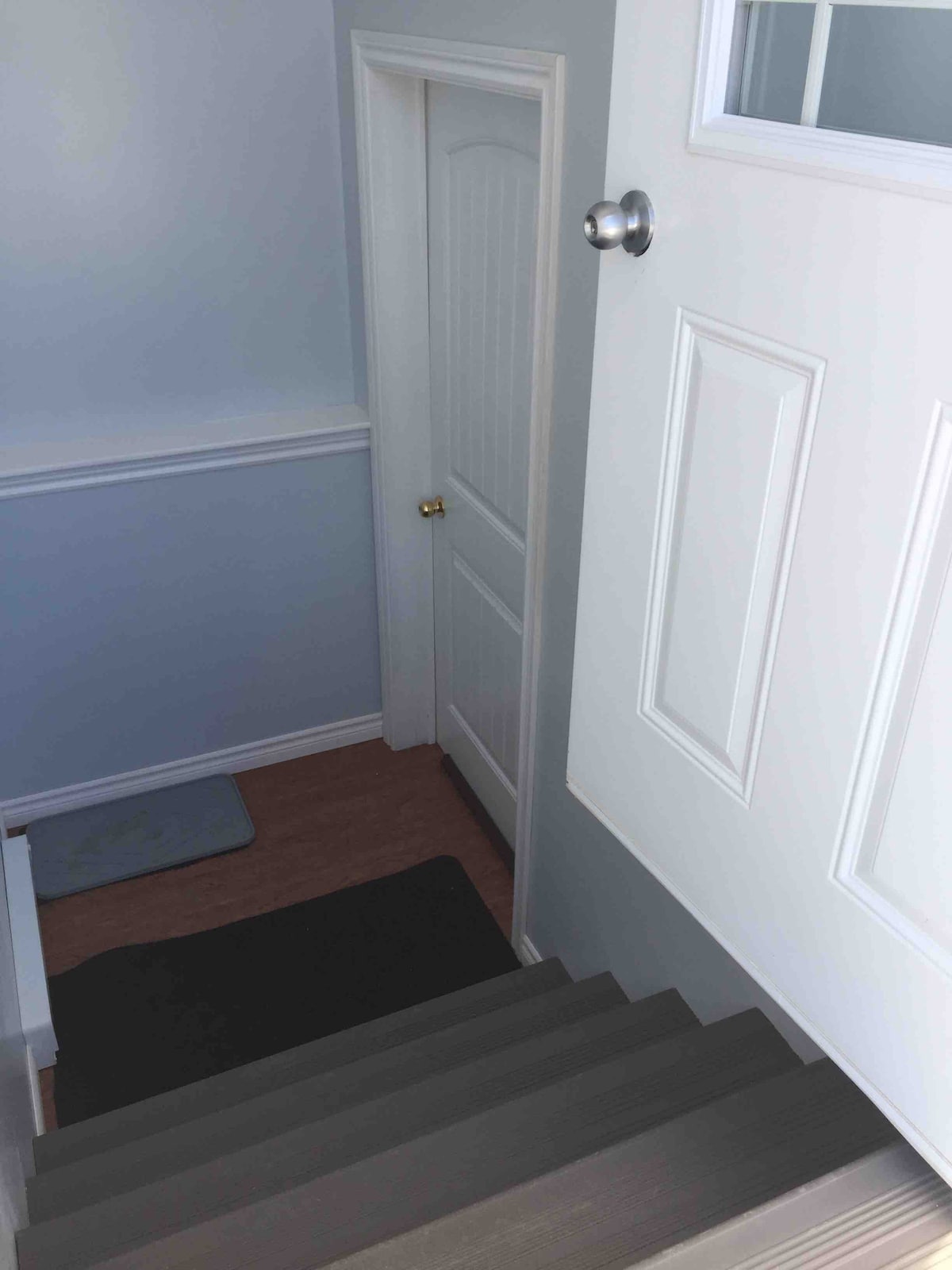A view of the staircase leading down is presented, with light beige steps and a white railing. At the bottom, a grey mat is placed on the floor beside a closed white door that features a round doorknob. Soft wall colors create a calm environment.