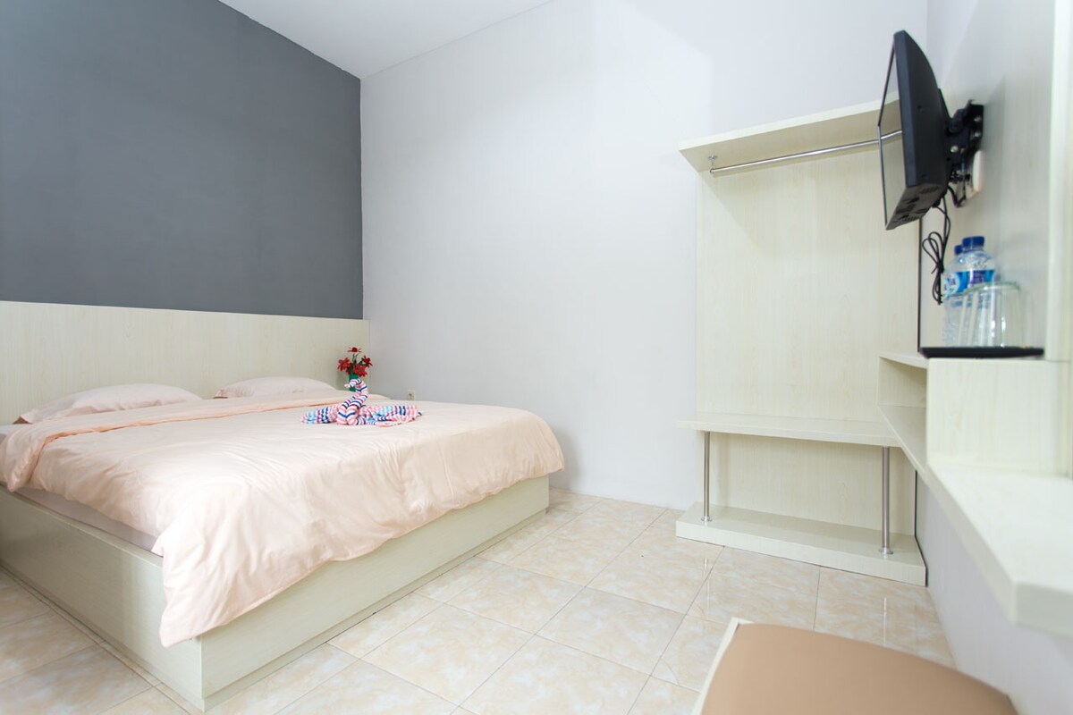 The room is furnished with a spacious bed dressed in light-colored linens. A mounted flat-screen TV is positioned on the wall opposite the bed. A simple shelving unit provides additional storage, while a small chair is placed nearby. Natural light brightens the neutral-toned space.