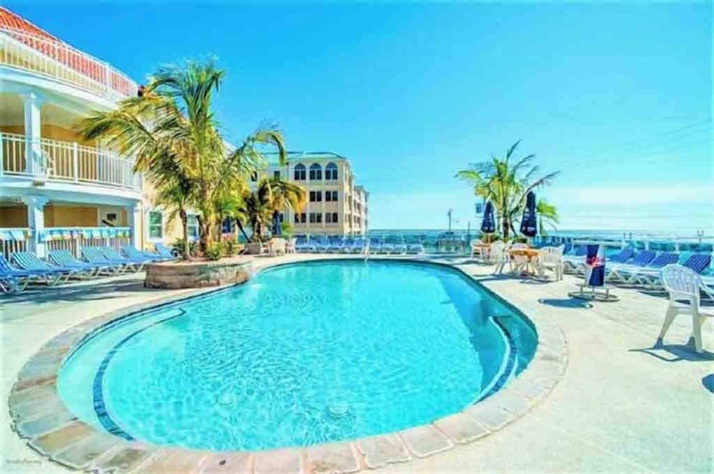 An inviting outdoor pool area features a clear blue water surface, surrounded by lounge chairs and tropical palm trees. The pool's shape is irregular, providing a relaxed setting for sunbathing and leisure. Nearby, a spacious deck extends with additional seating options under a bright sky.