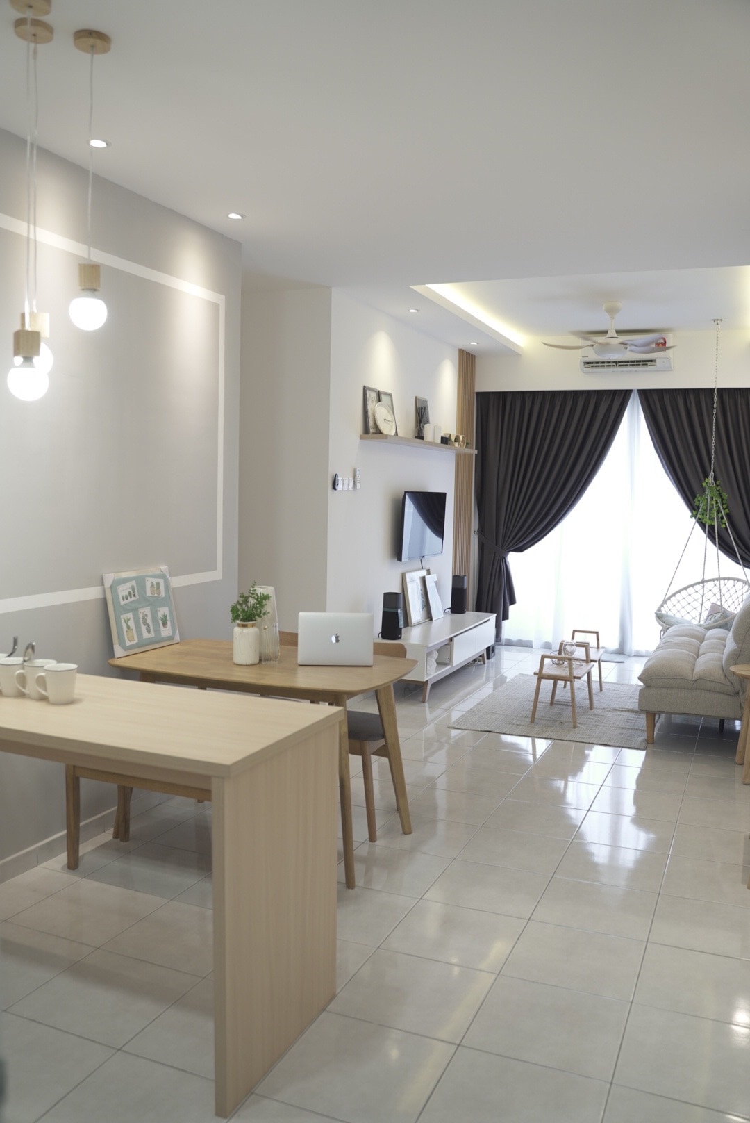 A modern living area features glossy tiled flooring and soft neutral walls. A light wood dining table with minimalist decor is present, alongside a comfortable sofa set. Natural light floods the space through a large window, complemented by sheer drapes and a decorative indoor plant.
