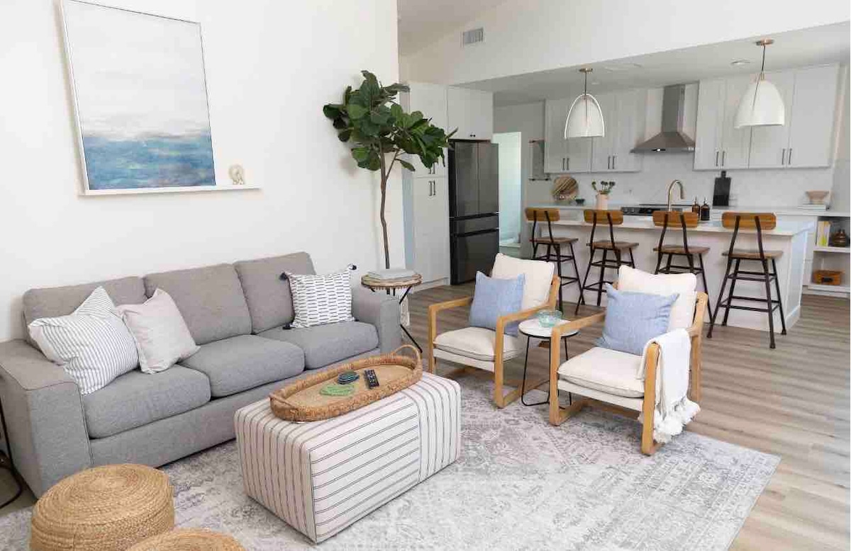 A modern living area features a comfortable gray sofa with decorative cushions, accompanied by two wooden chairs with blue accents. A coffee table with a striped design is placed on a textured area rug. The open plan kitchen is visible in the background, with bar stools and stainless-steel appliances.