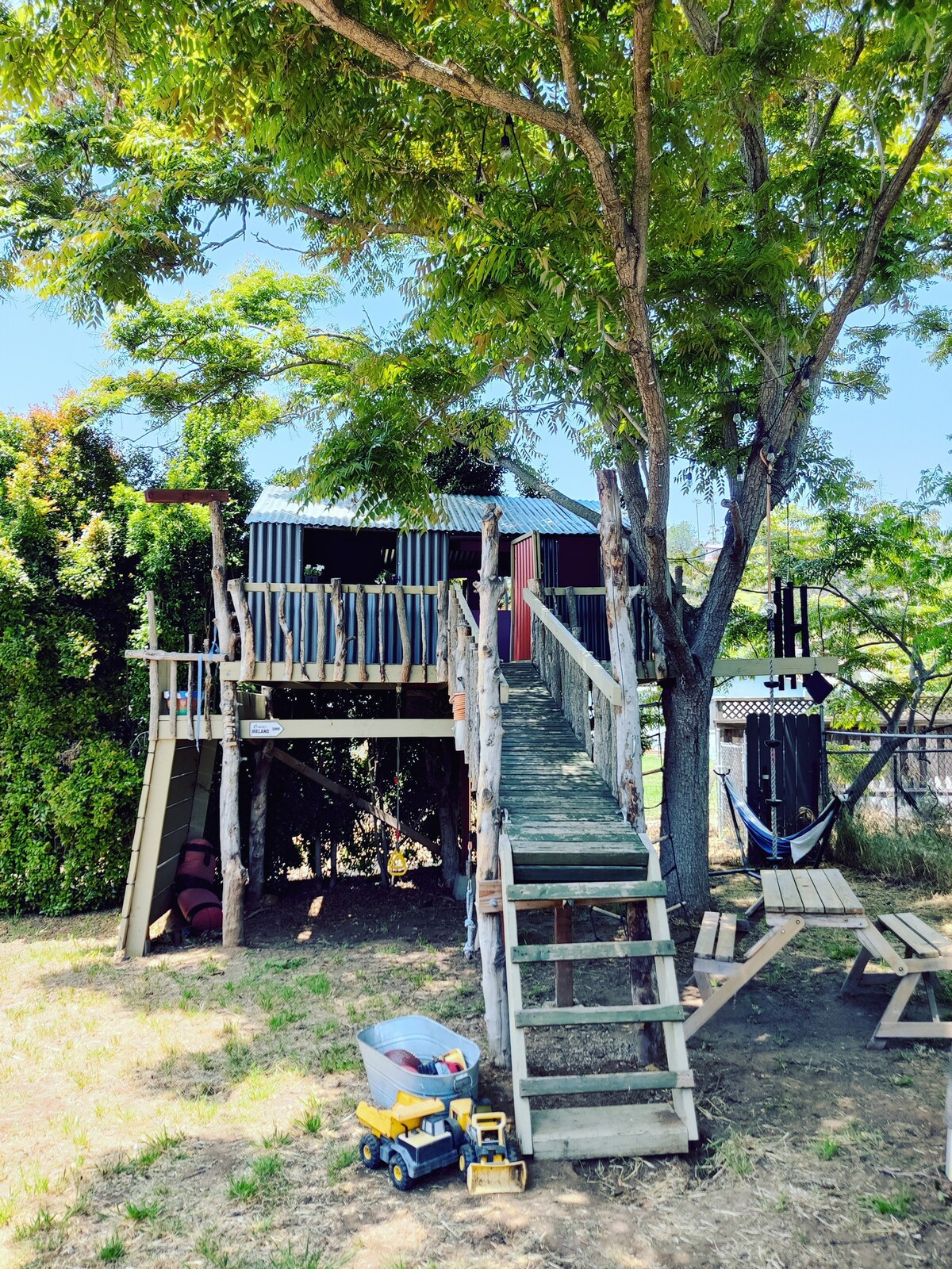 A wooden treehouse is perched among leafy trees, accessed by a sturdy staircase. Nearby, a shaded area features a hammock and benches, while a small toy truck rests on the ground, suggesting a playful environment for children.