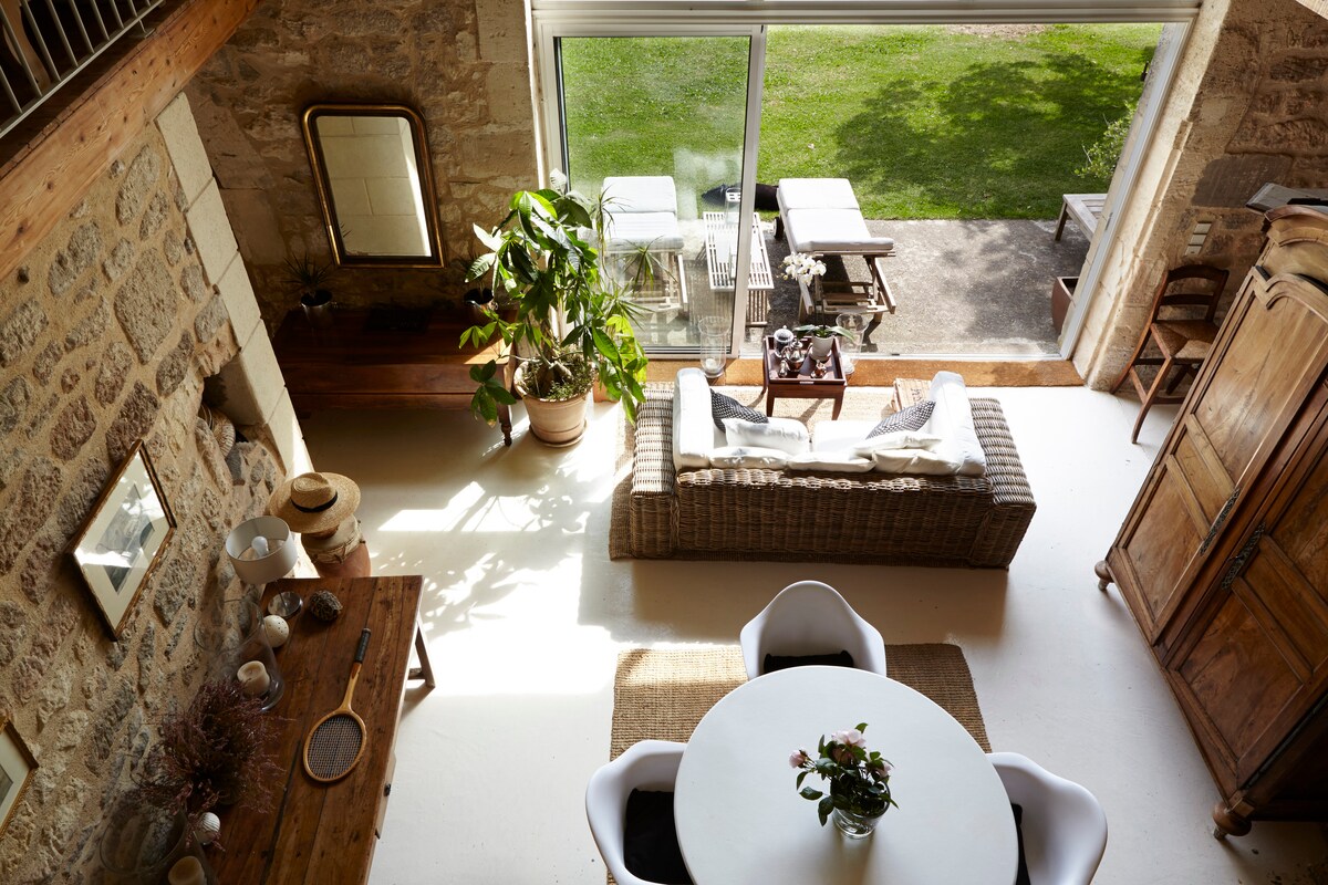 An inviting living area features a large glass door that opens to a green outdoor space. The interior is highlighted by a cozy seating arrangement with a woven sofa and a round dining table adorned with a floral centerpiece. Natural light floods the space, enhancing the rustic stone walls.