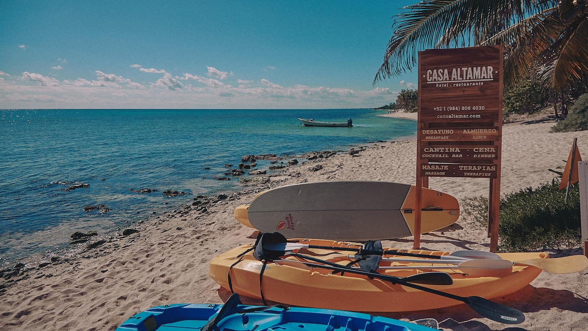 A sandy beach is viewed with calm waters stretching into the distance. Colorful kayaks are arranged near a wooden sign identifying 'Casa Altamar.' Tropical foliage frames the scene, while a small boat is anchored offshore under a clear blue sky.