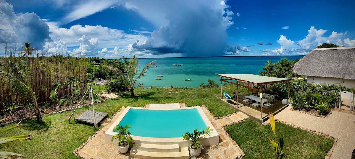 A clear view of the private swimming pool is presented, framed by lush greenery and a beachfront pavilion. The ocean glistens in the distance under a partly cloudy sky, providing a serene backdrop for relaxation. The surrounding landscape includes gentle slopes and a sandy area.