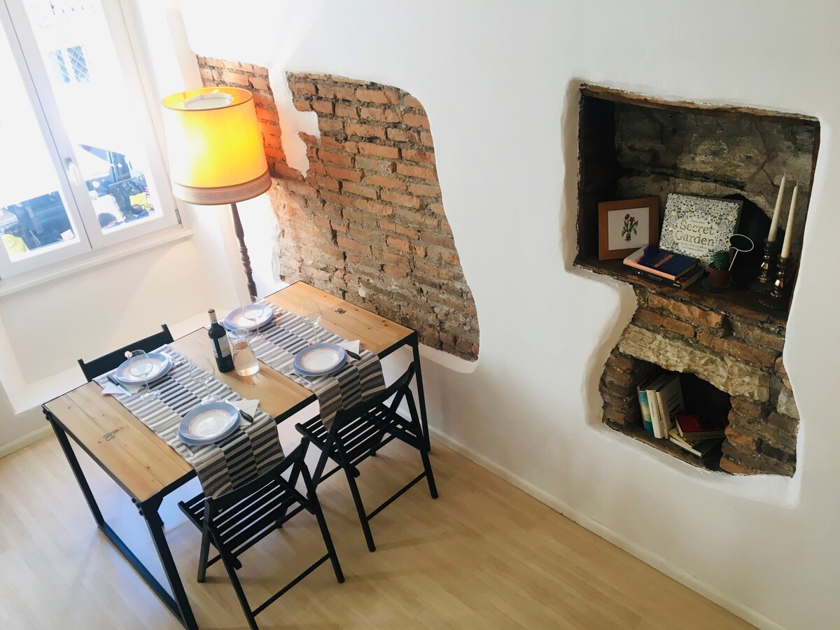 A dining area is presented with a wooden table surrounded by black chairs, set for four. Exposed brick walls add texture, complemented by a lamp that provides soft illumination. A small alcove displays books and candles, enhancing the inviting atmosphere.