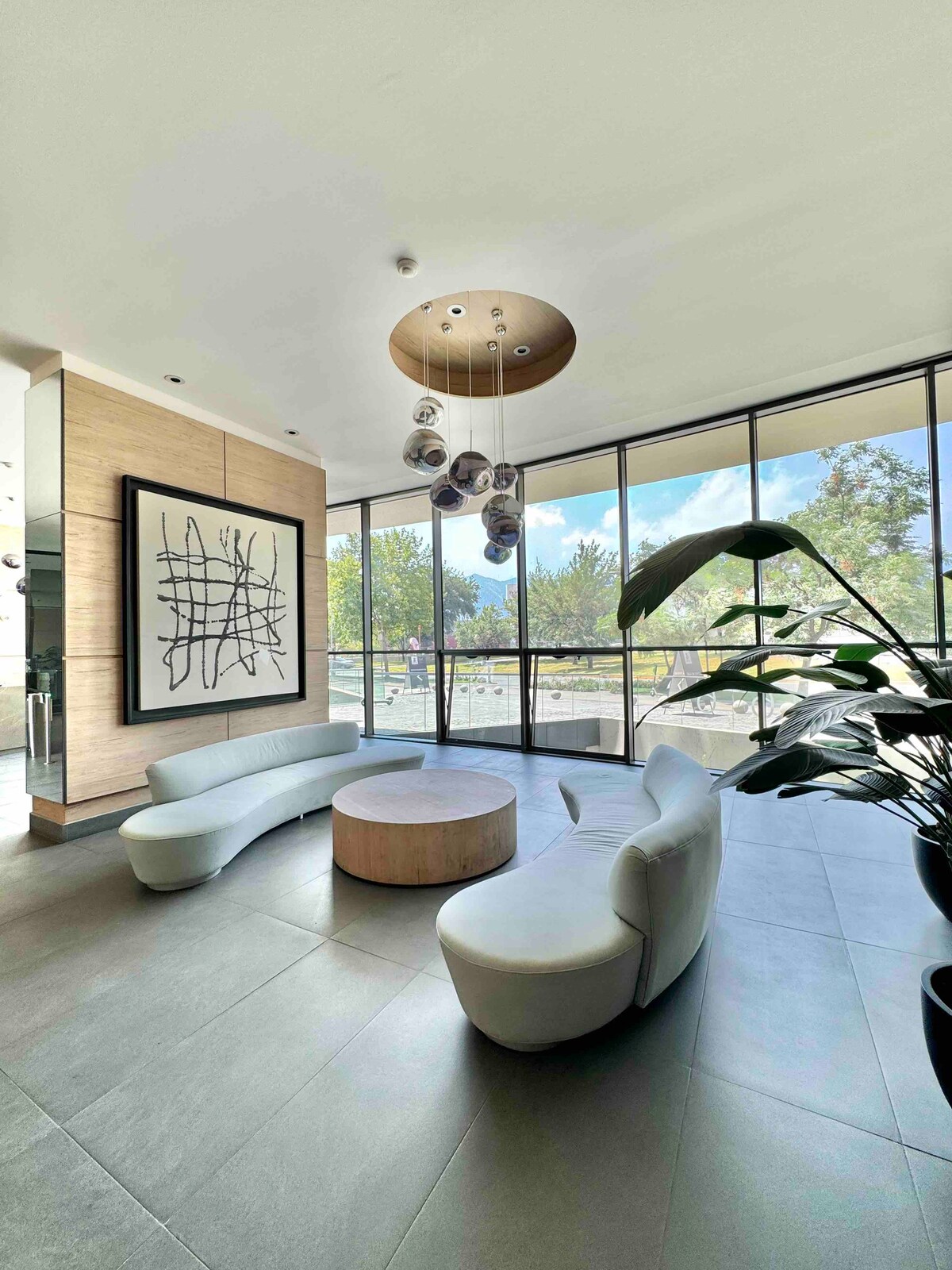 A modern lobby area is presented with large floor-to-ceiling windows offering views of surrounding greenery. Sleek, curved white seating is arranged around a circular wooden table, with a statement piece of artwork on the wall. A decorative light fixture hangs above, adding a contemporary touch.