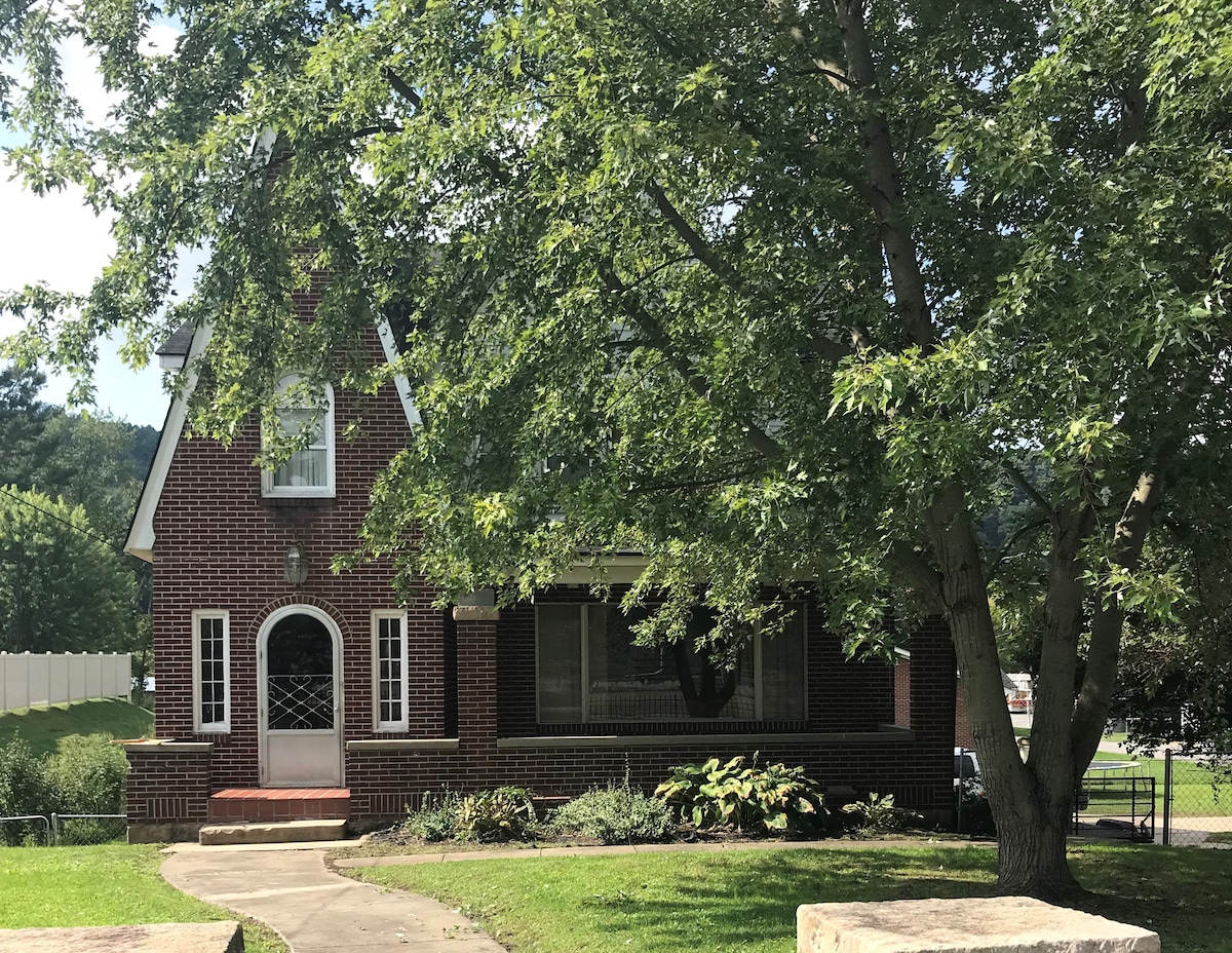 A charming brick house is set within a green yard, shaded by large trees. A welcoming entrance features a decorative front door and a small set of stairs leading to a well-maintained porch. Lush plants surround the base, enhancing the homey atmosphere.