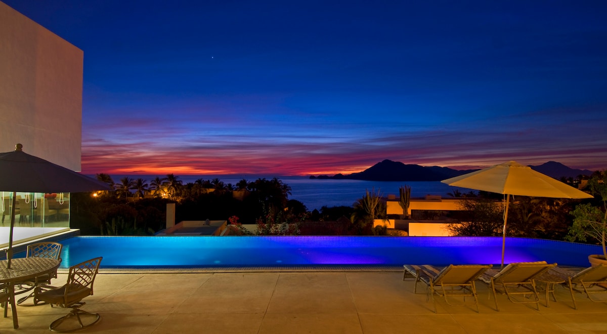 A serene infinity pool is highlighted against a vibrant sunset, showcasing hues of purple and orange over the Pacific Ocean. Comfortable lounge chairs line the poolside, complemented by a dining table beneath an umbrella, offering a tranquil space for relaxation.