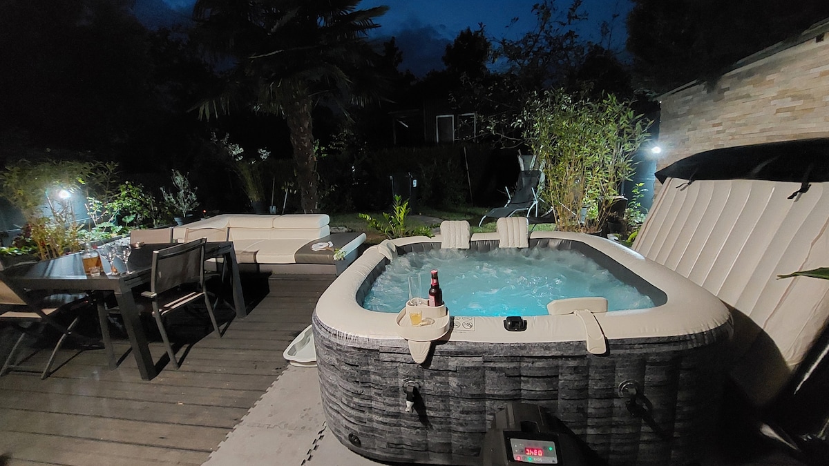 A private outdoor area features a jetted jacuzzi surrounded by tropical plants. Comfortable seating is arranged nearby, with a dining table set for outdoor meals. Soft lighting creates a serene atmosphere, enhancing the inviting space for relaxation.