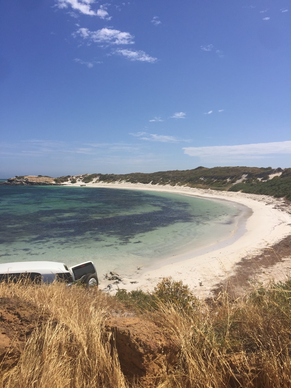 A serene coastal scene features a gentle curve of sandy beach meeting clear turquoise waters. Lush greenery borders the beach, providing a natural backdrop. Light clouds drift across a bright blue sky, creating a peaceful atmosphere. A parked van is visible near the beach's edge.