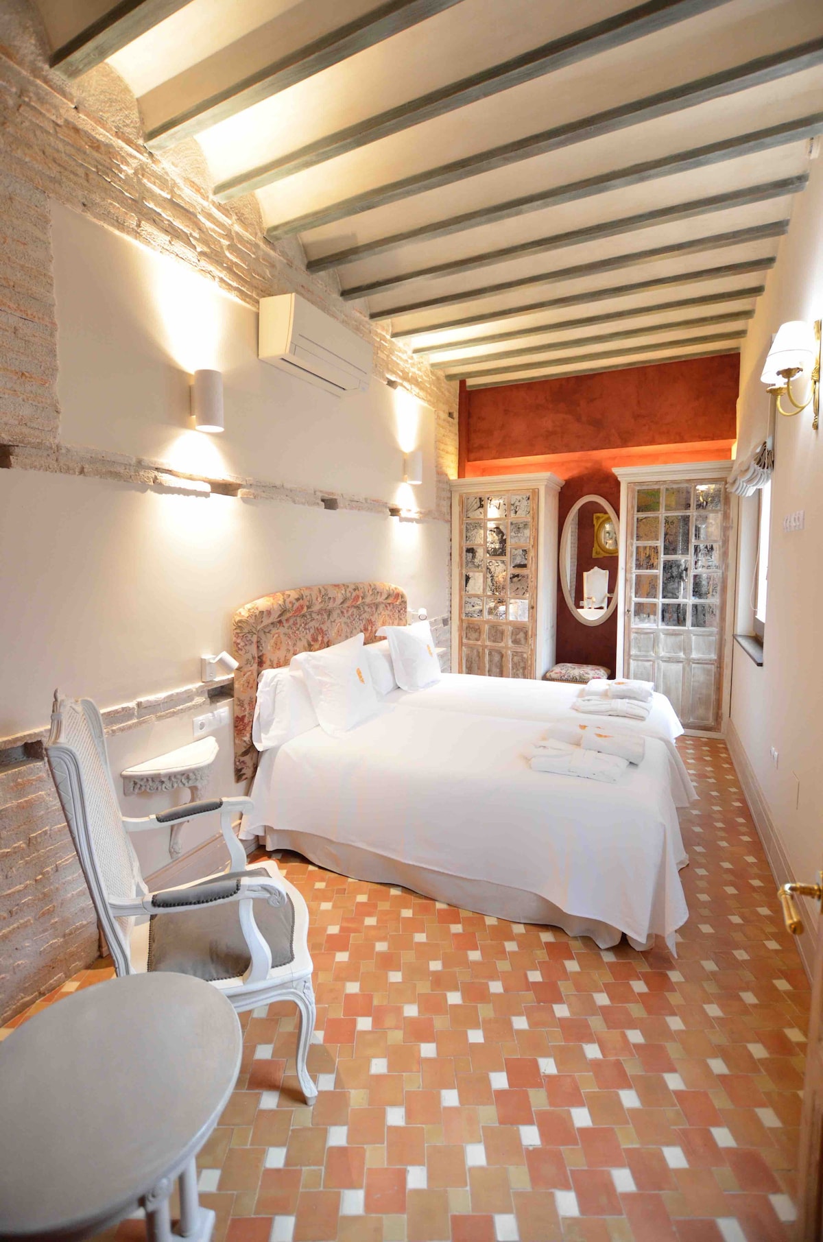 A spacious sleeping area features two large beds dressed in crisp white linens, complemented by neatly arranged towels. Natural light enters through delicate double doors, illuminating the room's elegant decor with exposed brick walls and warm, earthy tones.