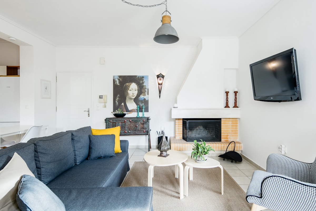 A spacious living area features a comfortable dark sofa, accented with a bright yellow pillow. A coffee table sits in the center, surrounded by stylish accent chairs. A wall-mounted television and a decorative fireplace add functional and aesthetic elements to the neutral decor.