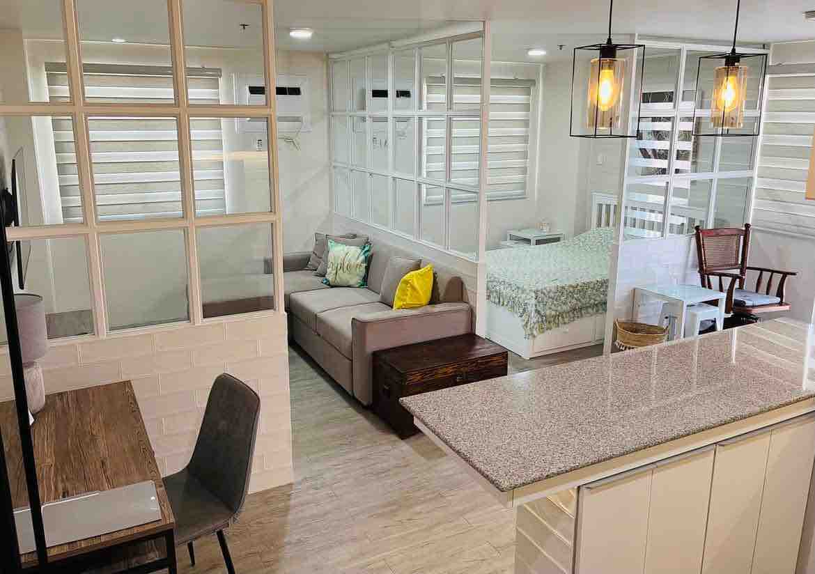 A bright living space is divided into areas for relaxation and sleeping. The couch is complemented by colorful cushions, and a small work desk is positioned nearby. Natural light filters in through the glass-paneled wall, illuminating the comfort of the shared areas.