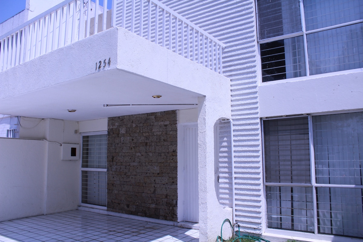 A modern entrance is seen, featuring a combination of textured stone and smooth white walls. Large windows allow ample natural light to enter. A covered parking area is visible, providing convenient access to the entrance of the property.