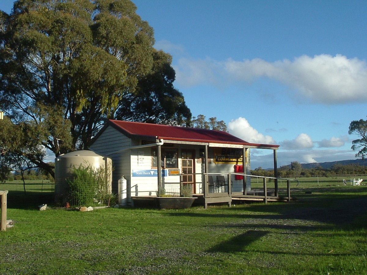 A charming cabin with a red roof is situated on a grassy plot, surrounded by trees and open fields. A large water tank stands beside the structure, which features a welcoming porch and signage. Mountains are visible in the distance under a partly cloudy sky.