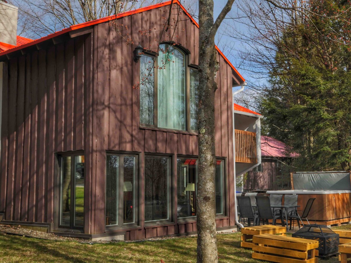 The exterior of the chalet features a modern design with large windows allowing ample natural light. A spacious patio area includes seating and is positioned near the spa, surrounded by grassy landscaping and trees.