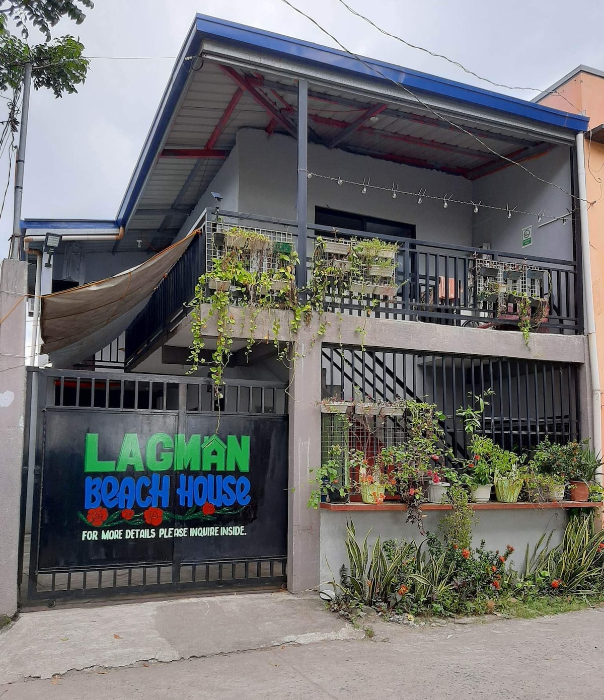 The exterior of Lagman Beach House is presented, featuring a two-story structure with a covered entrance. Green plants are arranged along the walls, and a decorative sign displays 'Lagman Beach House.' An inviting stairway leads to the upper level, complementing the overall design.
