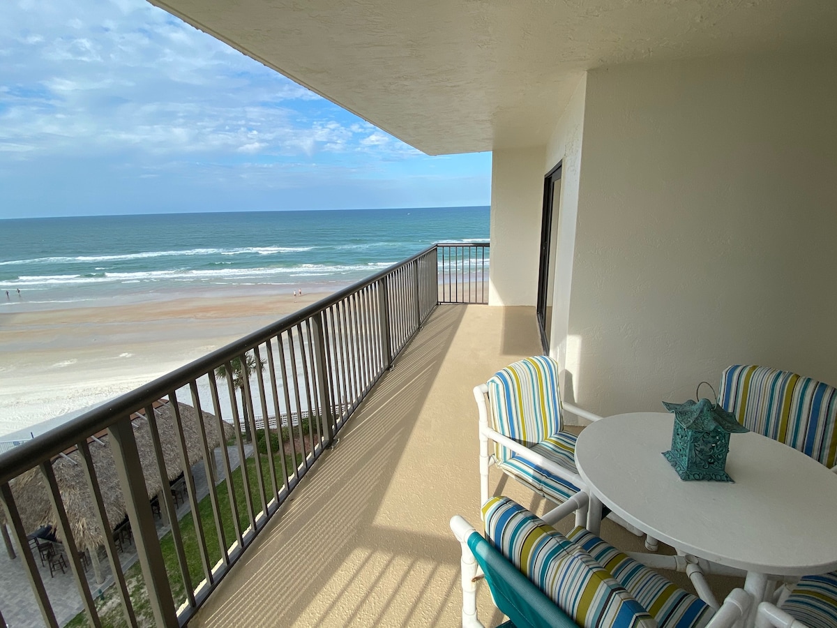 A spacious balcony offers stunning ocean views and features a round table with light-colored chairs adorned in striped patterns. The shoreline is visible below, with sand meeting the waves. The area is bathed in natural light, creating a serene coastal atmosphere.