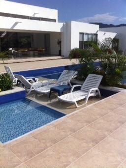 A serene outdoor area features two swimming pools surrounded by modern lounge chairs. Lush greenery and palm plants add a touch of nature, while the white exterior of the house provides a spacious backdrop against a clear blue sky.
