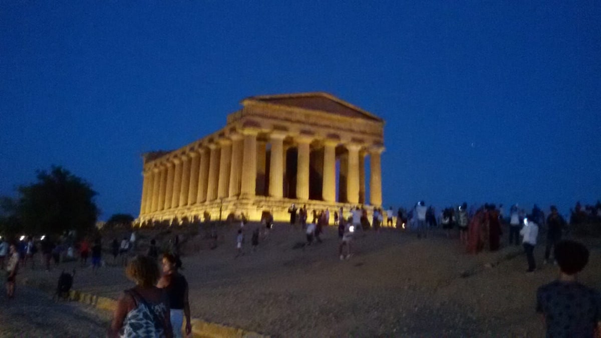 An ancient temple structure is illuminated against a twilight sky, highlighting its grand columns and architectural details. Visitors are seen exploring the area, enhancing the lively atmosphere surrounding the site. The surrounding landscape features uneven terrain leading to the temple's base.