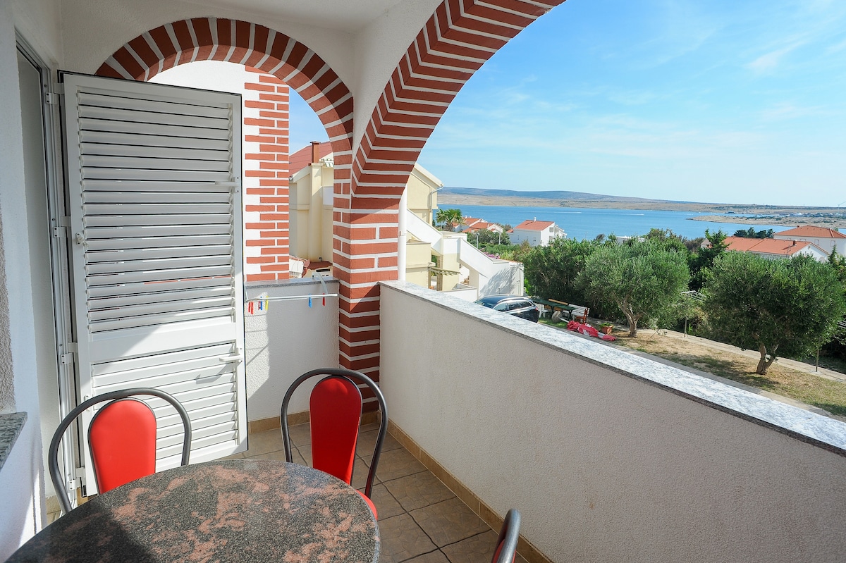 A view from a covered balcony features a round table with two red chairs, overlooking a landscape of greenery and water. The architecture includes distinctive red brick accents and a white exterior with a large arch framing the scenic vista.