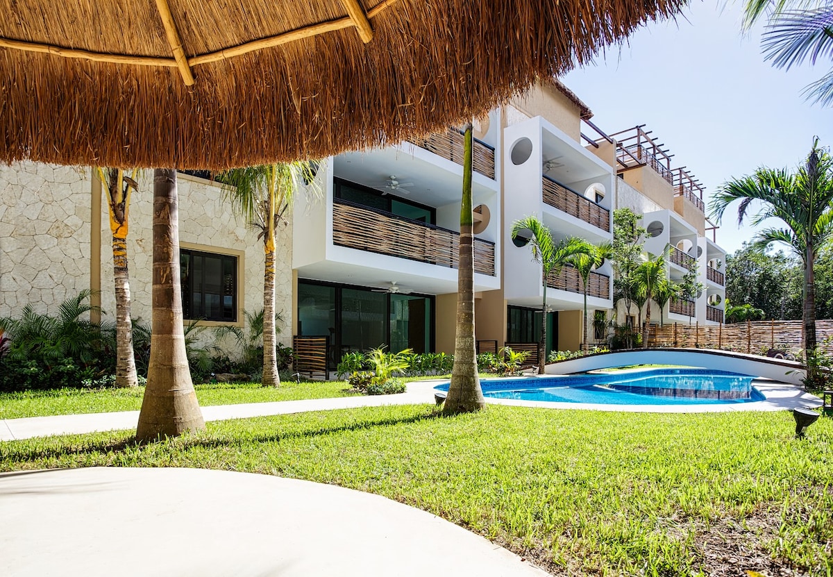 A landscaped area features lush green grass surrounding a swimming pool. The pool is framed by tropical plants and palm trees. White and beige buildings are visible in the background, showcasing multiple balconies with wooden railings under a clear blue sky.