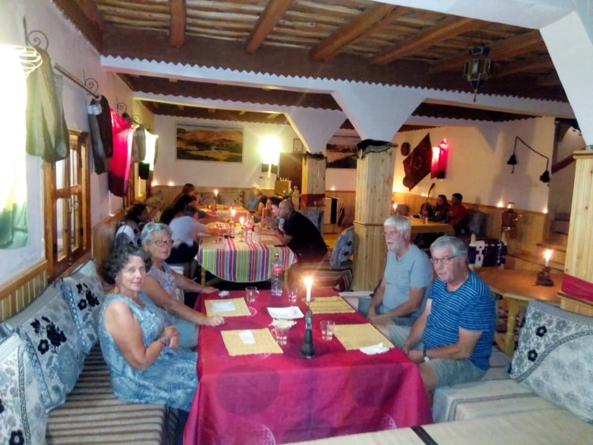 A dining area features comfortable seating arrangements with low tables, illuminated softly by candlelight and overhead fixtures. Colorful tablecloths adorn the tables, and guests are seen enjoying their meals in a warm, inviting setting. Wooden beams and traditional decor enhance the atmosphere.