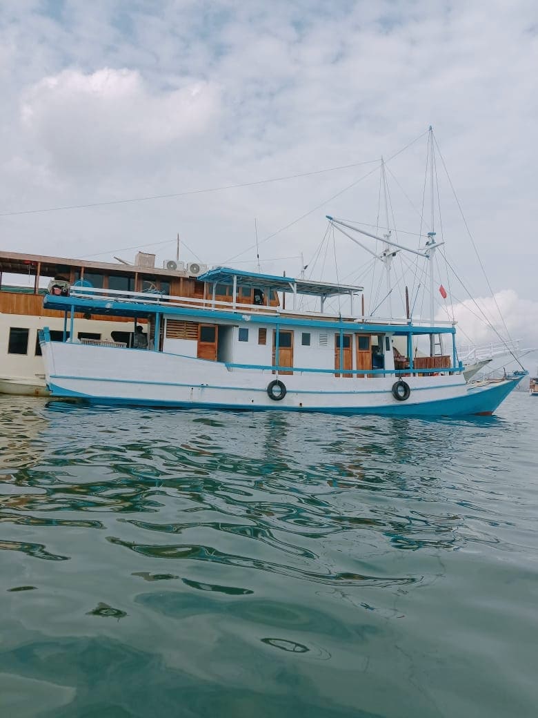 A traditional boat with a white hull and blue accents is anchored on calm waters. The vessel features multiple levels, with windows and an outdoor area visible. Scenic clouds can be seen in the sky, creating a serene backdrop for the boat.