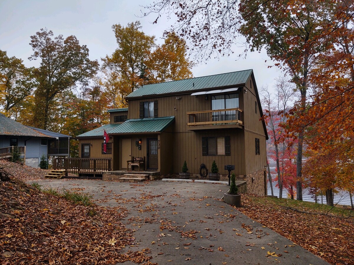 A two-story home is showcased with a metal roof and natural wood tones. The inviting front porch features seating and is surrounded by autumn leaves. Trees with vibrant fall foliage frame the property, with a glimpse of Watauga Lake visible in the background.