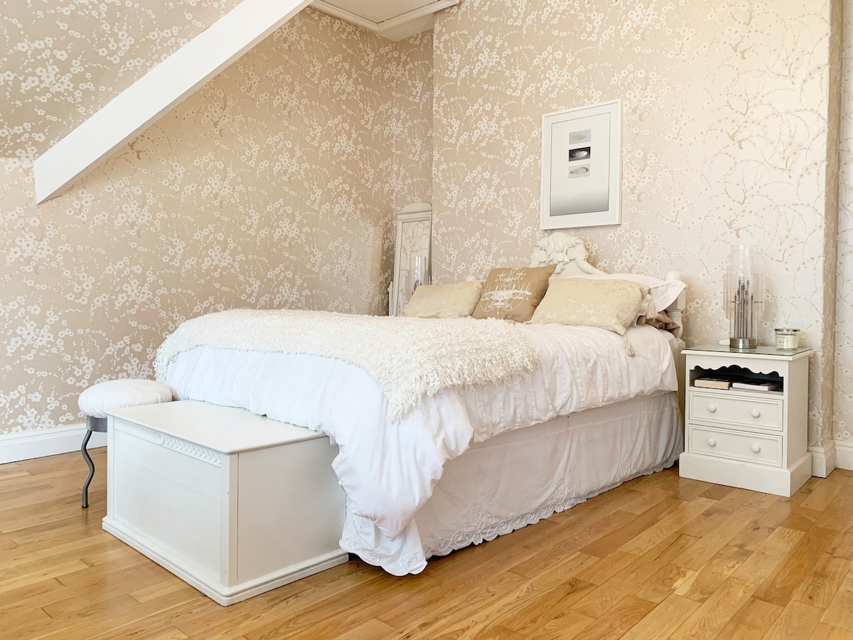 A spacious bedroom features a elegantly styled bed with a plush cream throw and various cushions on top. Light wooden flooring complements the soft floral wallpaper. A nightstand with drawers sits beside the bed, adding practical elegance to the inviting space.