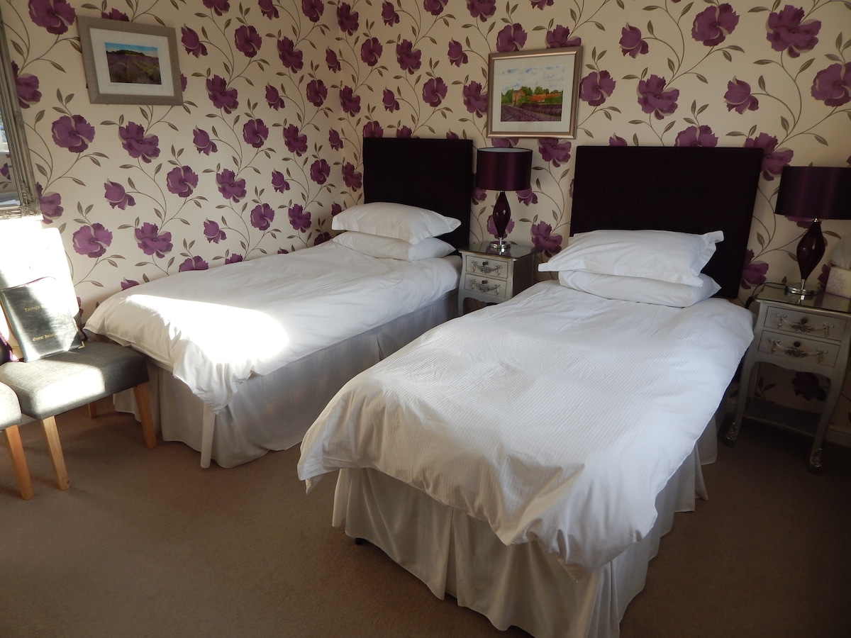 A well-appointed room displays two neatly arranged twin beds, each with white linens and plush headboards. Floral wallpaper in shades of purple adds a backdrop of color, while side tables with lamps provide soft lighting. A chair is positioned beside one bed, enhancing the room's functionality.