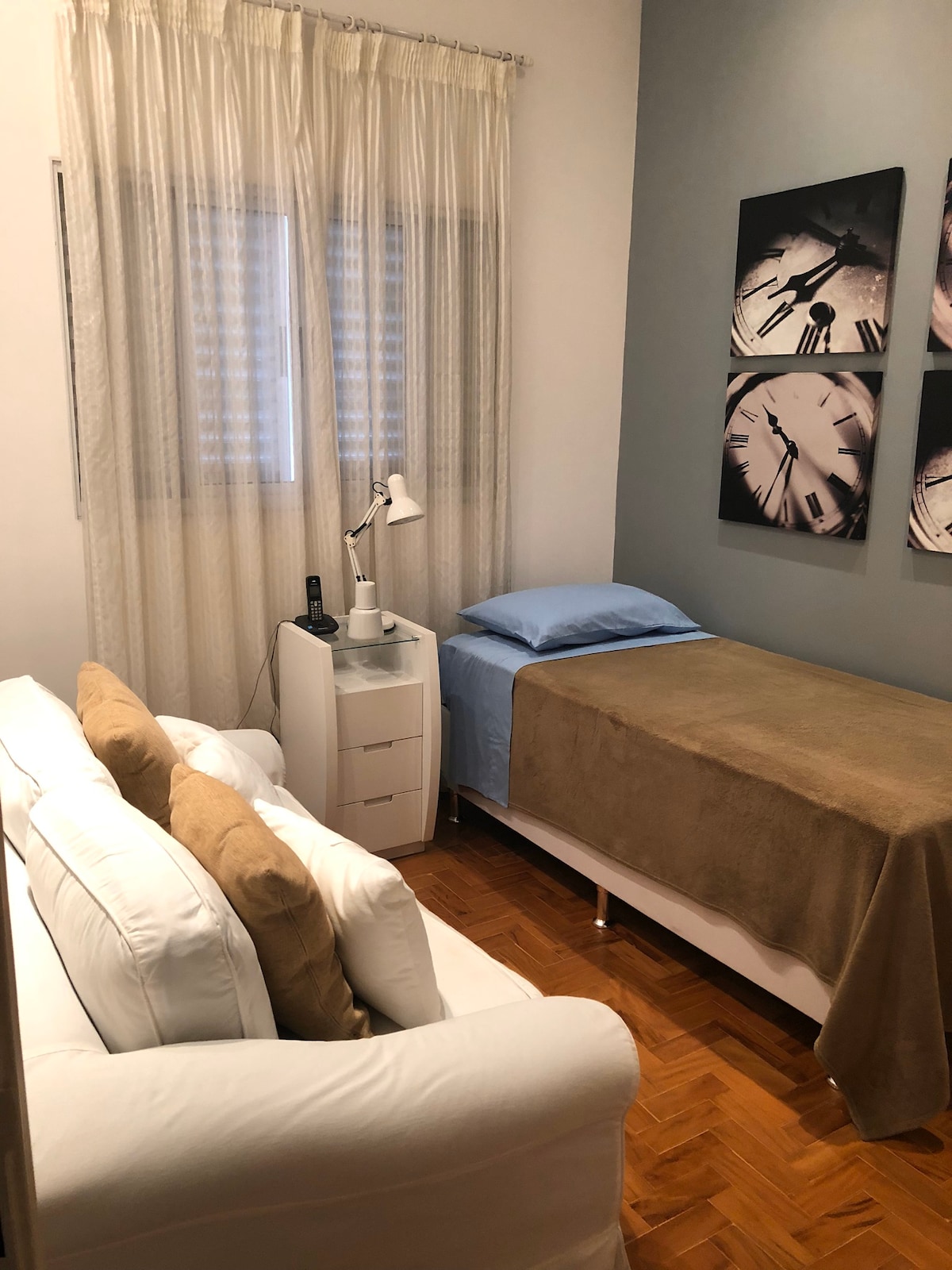 A compact, well-lit bedroom features a single bed covered with a brown blanket, positioned near a window draped in sheer curtains. A small bedside table sits beside the bed, complemented by a modern lamp. A white sofa with brown cushions occupies one corner of the room.