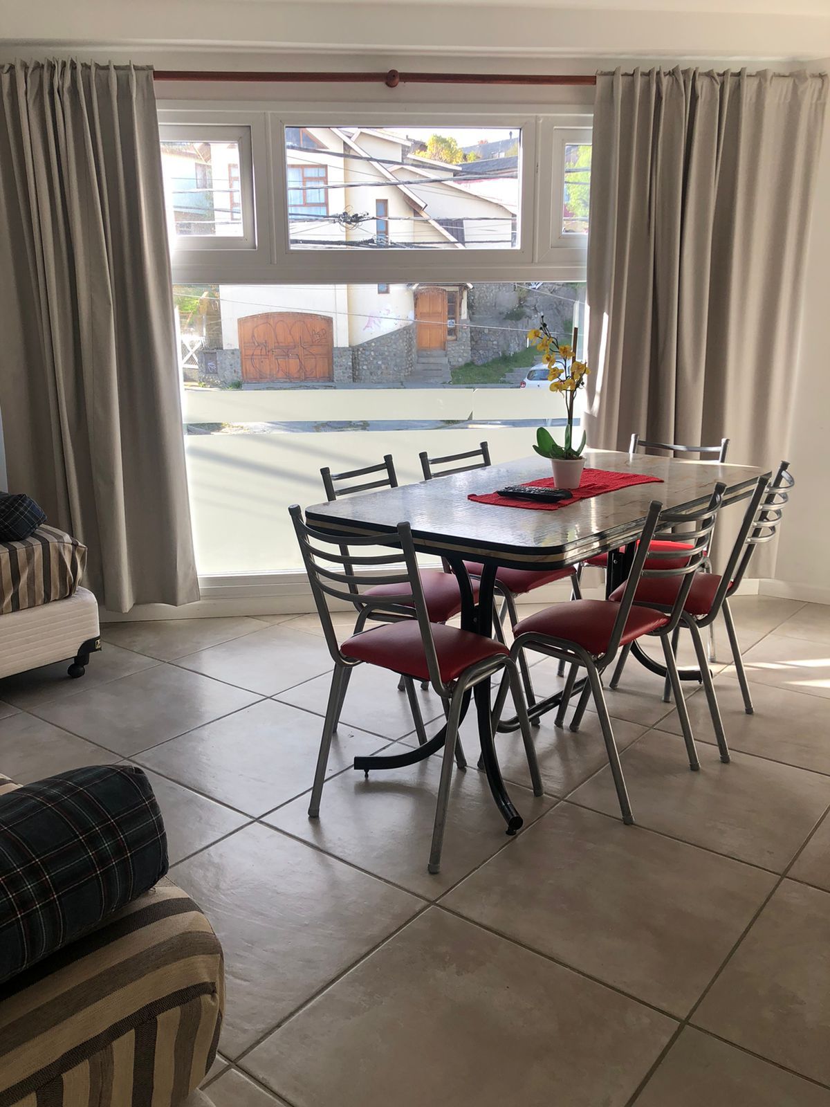 A dining area features a rectangular table surrounded by six metal chairs with red cushions. Natural light enters through large windows adorned with light curtains, revealing views of the outdoors. The tiled floor adds a modern touch to the space.