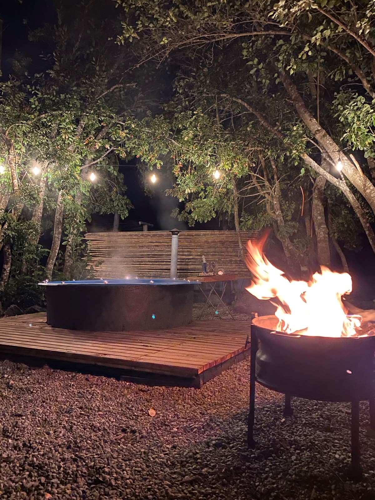 A private outdoor area features a round hot tub surrounded by trees, softly illuminated by hanging lights. Nearby, a fire pit displays warm flames, creating a cozy ambiance. The space is framed by gravel, providing a natural setting for relaxation.