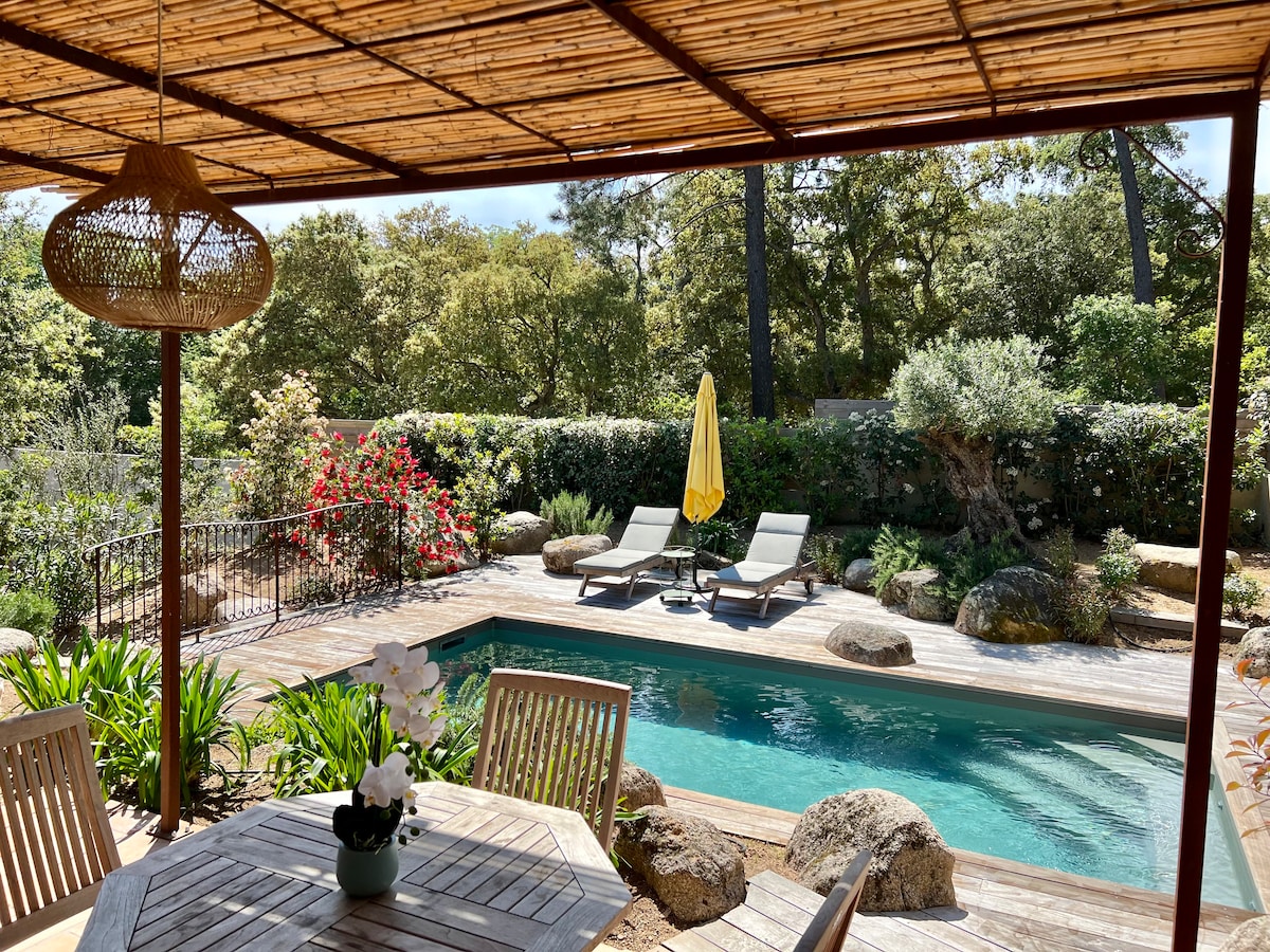 A serene outdoor space features a private heated swimming pool surrounded by lush greenery and decorative rocks. Sun loungers await relaxation under a yellow umbrella, while a wooden table and chairs provide seating nearby. Bright flowers add color to the tranquil garden setting.