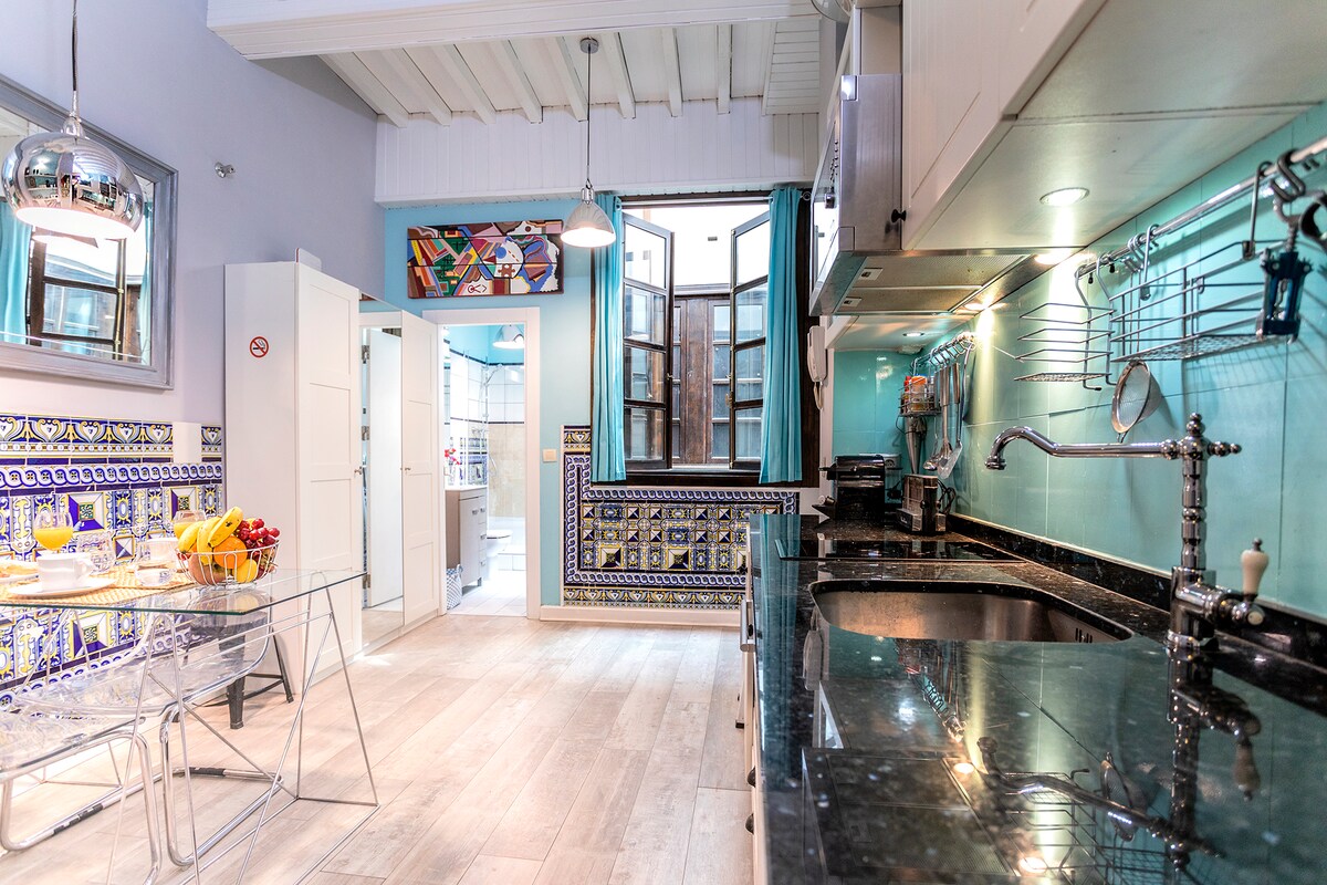 A modern kitchen features a black granite countertop, stainless steel fixtures, and colorful Spanish tiles. Natural light filters through open windows, illuminating a glass dining table accompanied by stylish chairs. The space is designed with efficient storage solutions and equipped with various kitchen appliances.
