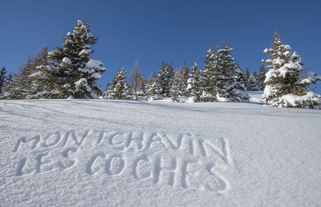 A snowy landscape is presented, with pine trees adorned in fresh snow. The words 'Montchavin Les Coches' are carved into the snow, creating a striking visual element against the pristine white background. The clear blue sky enhances the serene atmosphere.