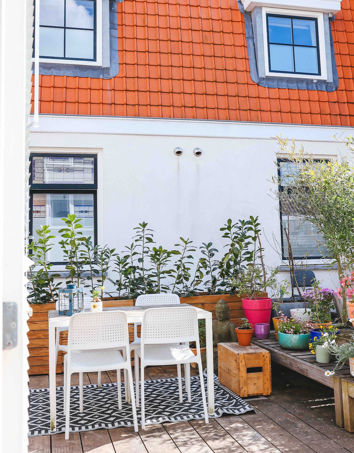 A charming outdoor patio is highlighted by a black and white geometric rug. White chairs surround a small table, complemented by a variety of potted plants that add greenery and color. Windows with dark trim can be seen in the background, enhancing the inviting space.