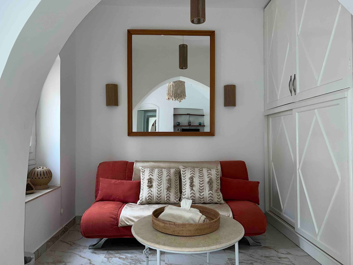 A cozy living area features a red sofa adorned with patterned cushions, complemented by a round coffee table. A large mirror above the sofa reflects the light, while wall sconces add soft illumination. Neutral tones and modern decor create a relaxed atmosphere.