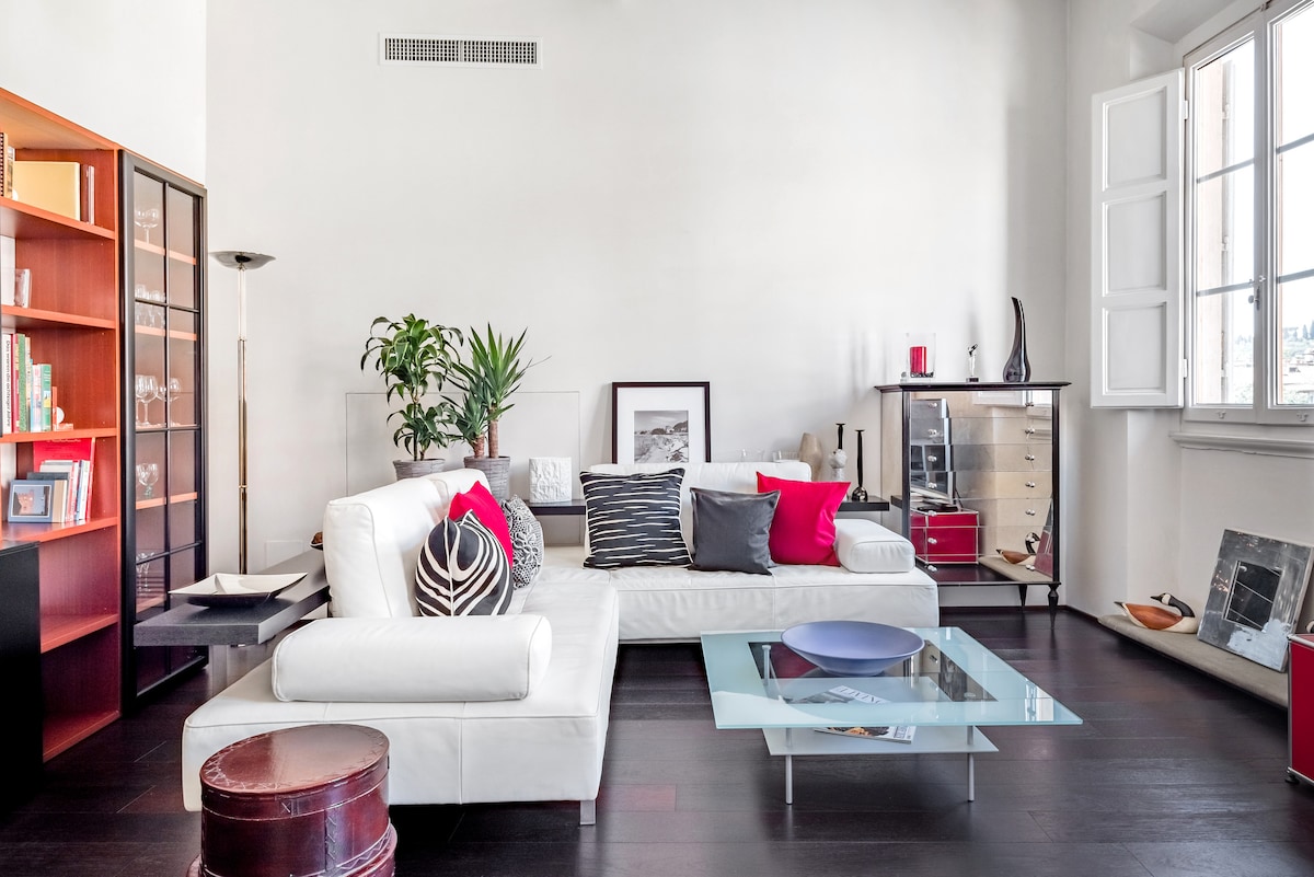 A modern living area features a white sectional sofa adorned with colorful pillows. A glass coffee table rests on dark hardwood flooring, surrounded by a tall bookshelf and a cabinet. Natural light enters through large windows with shutters, illuminating the space.